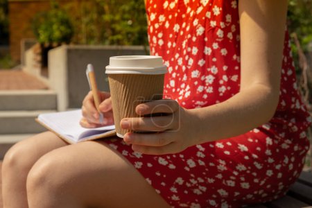 Photo for Unrecognizable Young woman in red dress drinking coffee from craft paper cup Writing Gratitude Journal on wooden bench. Today I am grateful for. Self discovery journal, self reflection creative writing, self growth personal development concept. Self - Royalty Free Image