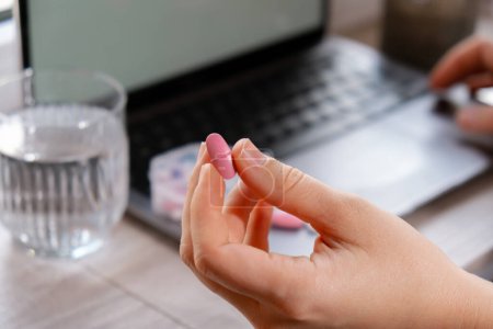 Frau, die täglich Vitamine weiß einnimmt, arbeitet am Laptop Organizer wöchentliche Aufnahmen Nahaufnahme der medizinischen Tablettenbox mit Tabletten für die tägliche Einnahme von Medikamenten mit weißen rosa Medikamenten und Kapseln. Arbeitsplatz