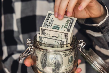 Foto de Dollar banknote saving money in glass jar. Unrecognizable woman moderate consumption and economy Collecting money. Tips. Business, finance, saving, banking and people concept. Extra money, passive - Imagen libre de derechos