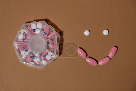 Foto de Organizador inyecciones semanales con sonrisa hechas de tabletas para el día a día tomar medicamentos con medicamentos de color rosa blanco y cápsulas. Vitaminas diarias en casa. Medicamentos: suplementos dietéticos, inmunidad y prevención de enfermedades - Imagen libre de derechos