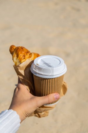 Photo for Tasty fresh croissants with coffee in paper cup in female hands on beach sea coast background. Breakfast outdoors wellbeing concept. FPV. Take away food. Coffee break alternatives - Royalty Free Image