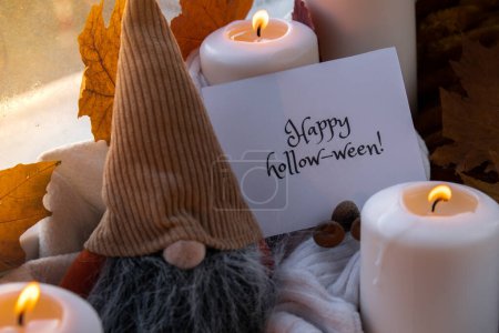 Foto de FELIZ HOLLOWEEN texto tarjeta de felicitación concepto Celebración de Halloween vacaciones de otoño en casa acogedora en el alféizar de la ventana Hygge ambiente estético hojas de otoño gnome especias y velas en blanco de punto - Imagen libre de derechos
