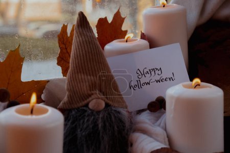 Foto de FELIZ HOLLOWEEN texto tarjeta de felicitación concepto Celebración de Halloween vacaciones de otoño en casa acogedora en el alféizar de la ventana Hygge ambiente estético hojas de otoño gnome especias y velas en blanco de punto - Imagen libre de derechos