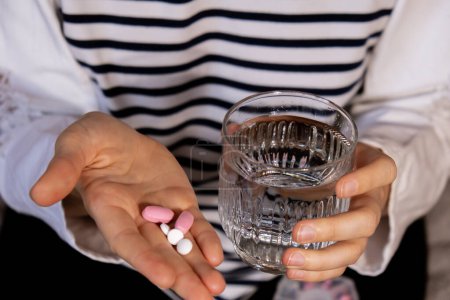 Photo for Female hands sorting pills Organizer weekly shots Glass of water Closeup of medical pill box with doses of tablets for daily take medicine with white pink drugs and capsules. Young woman getting her - Royalty Free Image