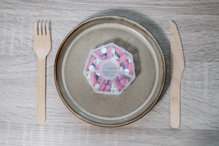 Téléchargez les photos : Organizer weekly shots with fork and knife medical pill box with doses of tablets for daily take medicine with white pink drugs and capsules. Daily vitamins at home. Medication dietary supplements - en image libre de droit