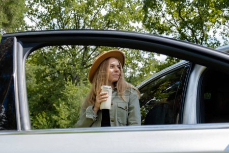 Photo for Blonde woman stoped on road next to car and drink coffee or tea from reusable mug. Refuse reuse recycle zero waste concept. Young tourist explore local travel making candid real moments. Responsible - Royalty Free Image