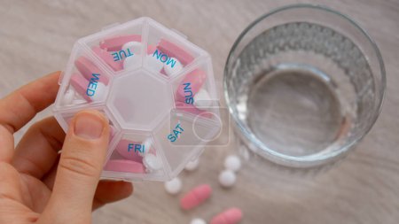 Téléchargez les photos : Woman sorting pills Organizer weekly shots Closeup of medical pill box with doses of tablets for daily take medicine with white pink drugs and capsules. Young woman getting her daily vitamins at home - en image libre de droit