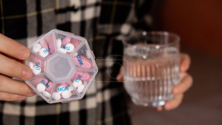 Téléchargez les photos : Female hands sorting pills Organizer weekly shots Glass of water Closeup of medical pill box with doses of tablets for daily take medicine with white pink drugs and capsules. Young woman getting her - en image libre de droit