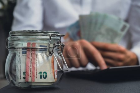Photo for Man hands counting expenses banknotes of euro cash from glass jar in the piggy bank on calculator. Close up of hands unrecognizable Businessman. Save up budget investment concept. Euros fund savings - Royalty Free Image