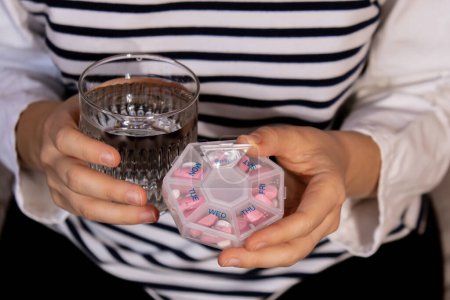 Photo for Female hands sorting pills Organizer weekly shots Glass of water Closeup of medical pill box with doses of tablets for daily take medicine with white pink drugs and capsules. Young woman getting her daily vitamins at home. Medication dietary suppleme - Royalty Free Image