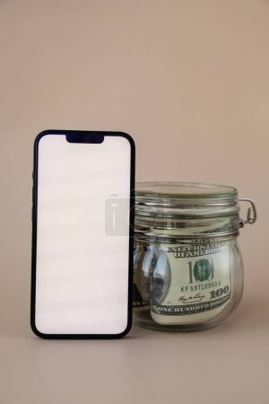 Photo for White smartphone screen mock up template in vertical position on beige background. Copy space App website advertising. Jar filled with dollars cash. Concept of Mobile application and technology - Royalty Free Image