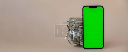 Photo for Banner Vertical Green screen on modern mobile phone in background of glass jar full of American currency dollar banknotes on beige background. Cope space for text. Advertisement for application - Royalty Free Image