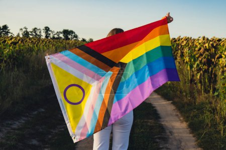 Foto de Mujer joven corriendo en el campo con la bandera Rainbow LGBTQIA ondeando en el viento hecho de material de seda sobre el fondo del campo. Símbolo del mes del orgullo LGBTQ. La igualdad de derechos. Concepto paz y libertad - Imagen libre de derechos
