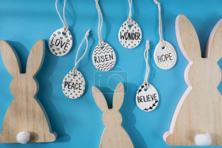 Group of wooden bunny ears and clay Easter eggs with words RISEN HOPE BELIEVE PEACE LOVE WONDER on blue background. Children air dry clay activity handicraft idea. Preparing for Easter holiday