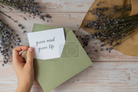 Female hands taking paper card note with text GREEN MIND GREEN LIFE from envelope. Lavender flower. Top view, flat lay. Concept of eco sustainability lifestyle mental spiritual health self care