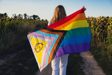 Symbol of LGBTQ pride month. Young woman showing Rainbow LGBTQIA flag waving in wind made from silk material on field background. Equal rights. Peace and freedom concept