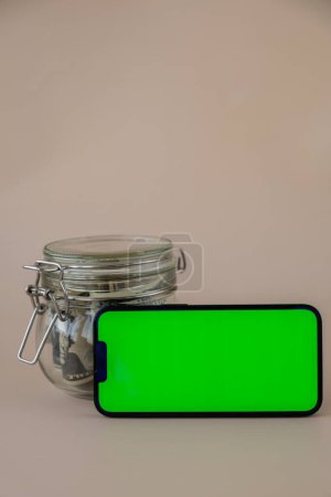 Chrome key smartphone screen mock up template in horizontal position on beige background. Copy space App website advertising. Jar filled with dollars cash. Concept of Mobile application and technology