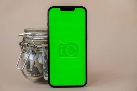 Vertical Green screen on modern mobile phone in background of glass jar full of American currency dollar banknotes on beige background. Cope space for text. Advertisement for application website