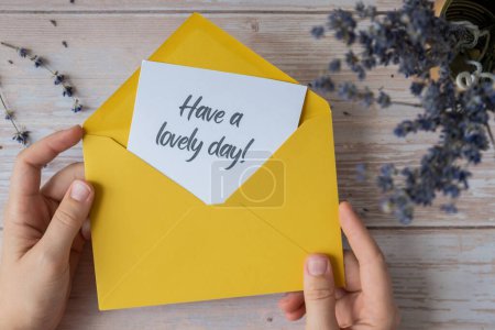 Female hands taking paper card note with text HAVE A LOVELY DAY from yellow envelope. Lavender flower. Top view, flat lay. Concept of mental spiritual health self care wellbeing mindfulness