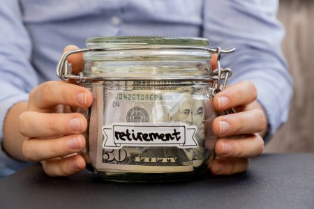 Unrecognizable woman holding Saving Money In Glass Jar filled with Dollars banknotes. RETIREMENT transcription in front of jar. Managing personal finances extra income for future insecurity background