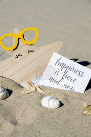 HAPPINESS IS HERE AND NOW text on paper greeting card on background of funny starfish in glasses summer vacation decor. Sandy beach sun coast. Slowing-down, enjoying the moment, good moments, slow life Holiday concept postcard. Getting away Travel Bu