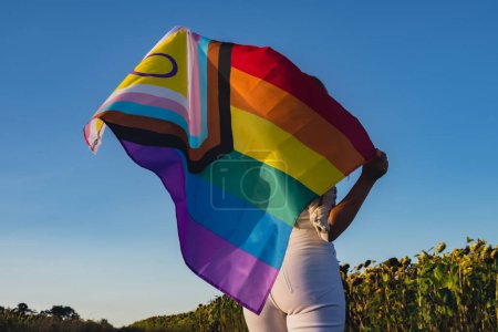 Girl showing her gender identity. Young woman stand with pride flag Rainbow LGBTQIA flag made from silk material on field background. Symbol of LGBTQ pride month. Equal rights. Peace and freedom