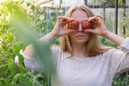 Porträt einer blonden Frau, die im Gewächshaus rote reife Bio-Tomaten erntet und Spaß hat. Gesundes hausgemachtes Nahrungsmittelkonzept. Landleben in Cottagecore 