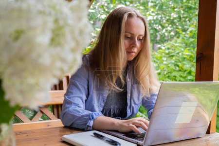 Étudiante a leçon en ligne éducation en plein air dans le jardin alcôve en bois. Femme blonde assise dehors travaille sur un ordinateur portable ayant un appel vidéo. Unité avec la nature