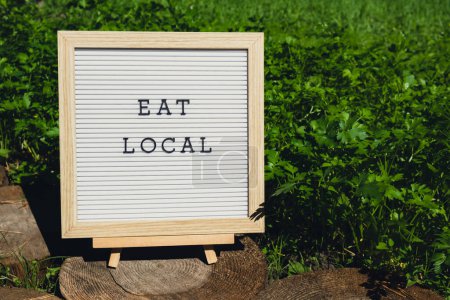 Letter Board mit Text EAT LOCAL auf dem Hintergrund des Gartenbeetes mit grünem Kraut Petersilie. Ökologischer Landbau, produzieren lokales Gemüse Konzept. Unterstützung lokaler Landwirte. Saisonmarkt 
