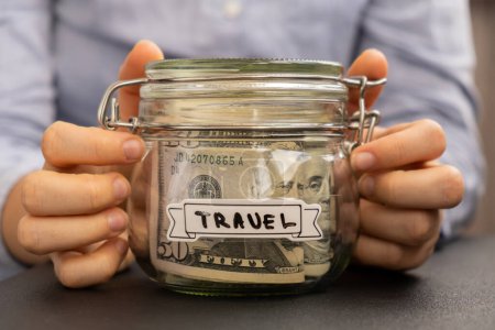 Unrecognizable woman holding Saving Money In Glass Jar filled with Dollars banknotes. TRAVEL transcription in front of jar. Managing personal finances extra income for future insecurity background