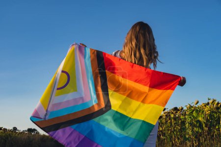 Symbol of LGBTQ pride month. Young woman showing Rainbow LGBTQIA flag waving in wind made from silk material on field background. Equal rights. Peace and freedom concept