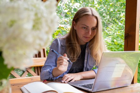 Female student has online lesson education outdoor in garden wooden alcove. Blonde woman sitting outside work on laptop having video call. Unity with nature