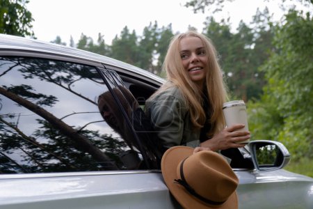 Smiling young woman looking from car window and drinking coffee or tea from reusable thermos cup. Local solo travel on weekends concept. Exited woman explore freedom outdoors in forest. Unity with
