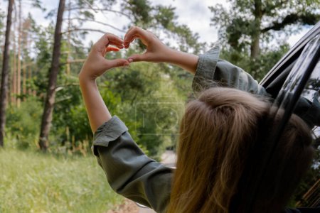 Woman riding car holding hand out of window showing heart symbol with her hands. Love and adventure Local traveling concept. Freedom and connection with nature. Sustainable lifestyle roadtrip on