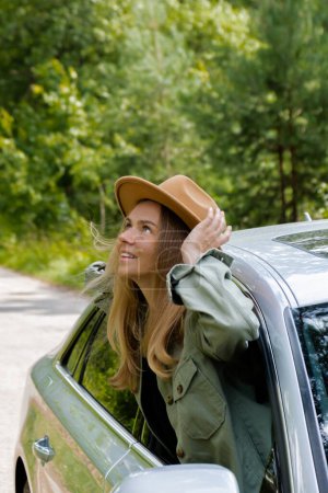 Blonde Frau mit Hut ragt Kopf aus Windschutzscheibe Auto. Junge Touristen erkunden die lokale Reisewelt und erleben echte Augenblicke. Wahre Emotionen, die sich an der frischen Luft entspannen und erfrischen