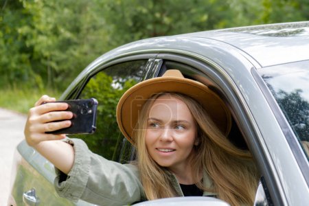 Aufgeregt lächelnde Frau macht Videoanruf mit Handy aus dem Autofenster. Lokale Alleinfahrten am Wochenende. Junge Reisende erkunden die Freiheit draußen im Wald und machen Selfie-Fotos. Einheit mit