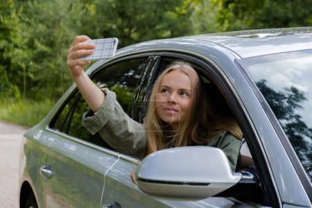 Blonde Frau stoppte Auto auf der Straße, um ein Selfie-Foto zu machen. Junge Touristen erkunden die lokale Reisewelt und erleben echte Augenblicke. Wahre Emotionen, die sich an der frischen Luft entspannen und erfrischen
