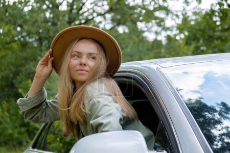 Lächelnde junge Frau mit Hut, die aus dem Autofenster schaut. Lokale Alleinfahrten am Wochenende. Aufgeregte Frau erkundet die Freiheit draußen im Wald. Einheit mit der Natur Lifestyle, Ruhe tanken Entspannung 