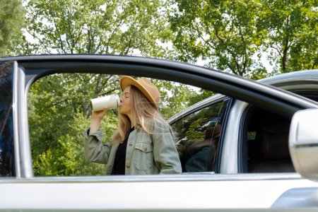 Blonde Frau hält auf der Straße neben dem Auto und trinkt Kaffee oder Tee aus Mehrwegbecher. Refuse Recycling Zero Waste Konzept. Junge Touristen erkunden die lokale Reisewelt und erleben echte Augenblicke. Verantwortlich