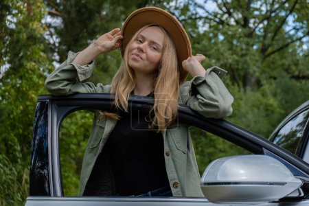Blonde woman in hat staying next to car door. Young tourist explore local travel making candid real moments. True emotions expressions of getting away and refresh relax on open clean air