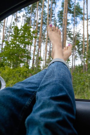 Weibliche Füße in blauen Jeans aus dem Autofenster. Konzept des komfortablen Reiseurlaubs. Wegkommen, um wirklich ehrliche Momente zu erleben. Reduzierung des CO2-Fußabdrucks. Frau rastet bei Roadtrip aus