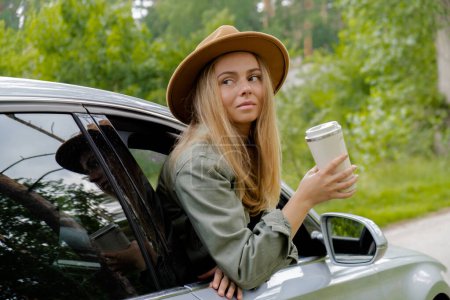 Blonde woman sticking head out of windshield car and drink coffee or tea from reusable mug. Young tourist explore local travel making candid real moments. Refuse reuse recycle zero waste concept