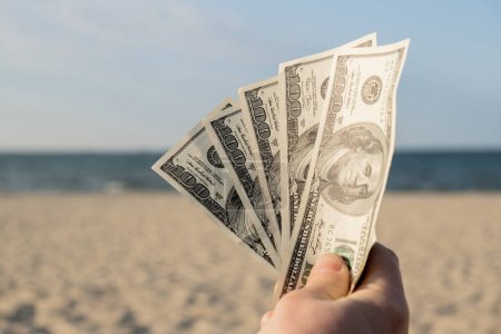 Unrecognizable woman Displaying Spread of Cash dollars bills on sandy beach coastline. Concept finance saving money for holiday vacation. Costs in travel holidays. Extra money, passive income