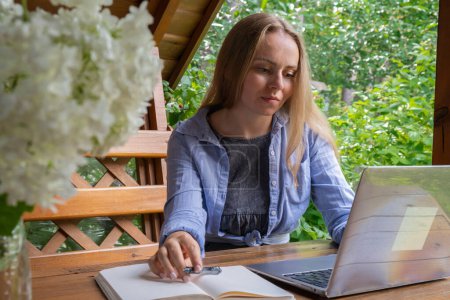 Estudiante tiene en línea lección de educación al aire libre en el jardín alcoba de madera. Mujer rubia sentada fuera del trabajo en el ordenador portátil teniendo videollamada. Unidad con la naturaleza
