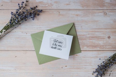 Paper card note with text SELF CARE IS THE NEW HEALTH CARE from green envelope. Lavender flower. Top view, flat lay. Concept of mental spiritual health self care wellbeing mindfulness