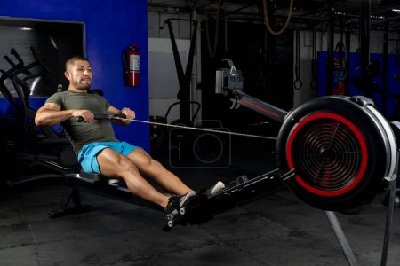 Photo for Side view of a Latin man with sportswear, rowing on a machine in a crossfit gym - Royalty Free Image
