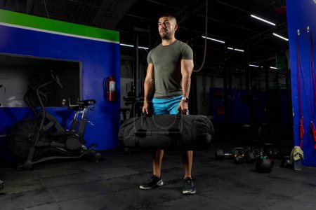 Photo for Male athlete wearing sportswear, standing in a gym holding a sandbag - Royalty Free Image