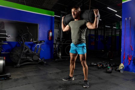 Photo for Latin man with sportswear, carrying a sandbag on his back while training in a gym - Royalty Free Image