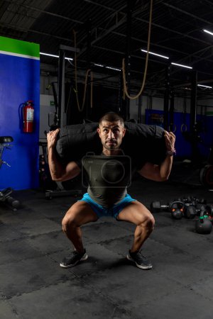 Photo for Man in sportswear in a gym doing squats with a sandbag on his back - Royalty Free Image