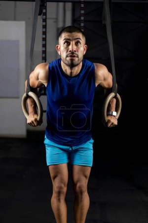 Photo for A Latino man in sportswear doing gymnastic exercises on some rings in a gym - Royalty Free Image